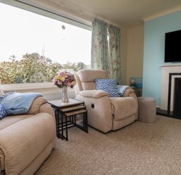 A living room with two beige armchairs a side table with flowers a fireplace and a large window with floral curtains at Rathillet Feock near Truro