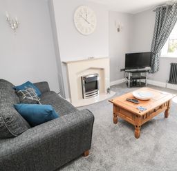 A living room with a grey sofa blue cushions a wooden coffee table a television on a stand a fireplace and a window with patterned curtains at Fisherman's Row in Amble