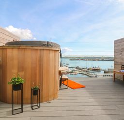 A balcony with a wooden hot tub two potted plants a wooden bench with cushions a small table with an orange rug and a view of boats on the water at 16 Arwenack Street in Falmouth