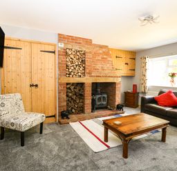 A living room with a brick fireplace stacked with firewood a wooden coffee table a leather sofa a patterned chair and a wall-mounted TV at Keepers Cottage in Beeston near Dereham