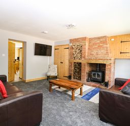 A living room with brown leather sofas red and gray pillows a wooden coffee table a brick fireplace and a wall mounted TV at Keepers Cottage in Beeston near Dereham