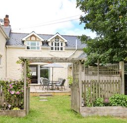 A garden with a wooden pergola and flower beds leading to a patio with a table, chairs, and an umbrella at Spring Cottage in Pentre-Cwrt