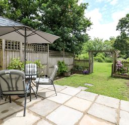 An outdoor patio with cushioned chairs around a table under an umbrella and a garden with a wooden fence and gate at Spring Cottage in Pentre-Cwrt