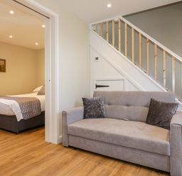 A room with a gray sofa under stairs and a bedroom with a bed visible through an open doorway at The Granary in Helland near Bodmin