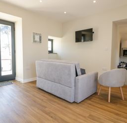 A living area with a gray sofa, a gray armchair, a wall-mounted TV, a clock, and a kitchen with appliances at The Hayloft in Helland near Bodmin
