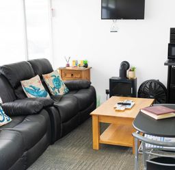 A living room with black leather sofas with sea creature cushions a wooden coffee table a wall-mounted TV a small wooden cabinet and a black table with chairs at 57 Cherry Park in Chapel St Leonards