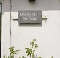 A black sign with white text saying Long Batch Cottage on a white wall between two windows at Long Batch Cottage in Ditcheat