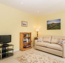A living room with a beige sofa a wooden bookshelf a floor lamp a small TV on a black stand and two pictures on yellow walls at Long Batch Cottage in Ditcheat