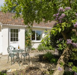A patio with a metal table and two chairs outside a cottage with a garden and flowering tree at Long Batch Cottage in Ditcheat