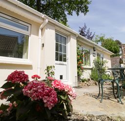 A patio area with pink flowers a white door windows and a green metal table and chairs at Long Batch Cottage in Ditcheat