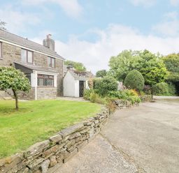 An exterior view of a stone farmhouse with a small leaning tree and raised garden bed on a lawn at South Boduel Farmhouse in Liskeard