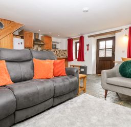 A living room with a gray sofa and orange cushions next to a kitchen with wooden cabinets at Keepers Cottage in Liskeard