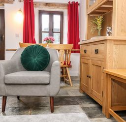 A room with a gray armchair with a green cushion a wooden cabinet a small wooden bench and a table with chairs near windows with red curtains at Keepers Cottage in Liskeard