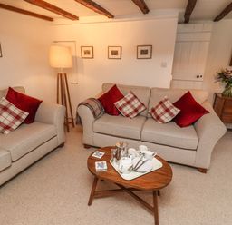 A living room with beige sofas and red and plaid cushions a wooden coffee table with tea set a floor lamp and a wooden chest with flowers at Lavender Cottage in South Zeal