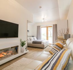 A living room with a fireplace a wall-mounted TV a beige sofa and a bed near glass doors at Westfields Kennards House near Launceston