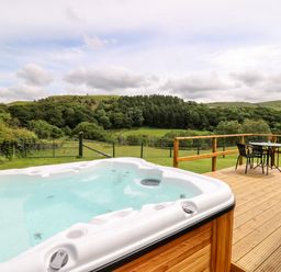 A deck with a hot tub and a table with chairs overlooking a grassy field and trees at Hwyl Rhayader near Llandrindod Wells