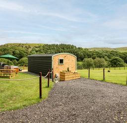 A small wooden cabin with a gravel path and picnic table with umbrella in a green field at Hwyl Rhayader near Llandrindod Wells