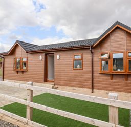 An exterior view of a brown wooden house with multiple windows a door and a small fenced area with artificial grass at 12 Bucklesham Park in Bucklesham near Trimley St Martin