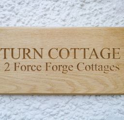 A wooden sign engraved with Turn Cottage 2 Force Forge Cottages on a textured white wall at Turn Cottage in Satterthwaite near Hawkshead