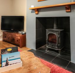 A wood burning stove in a fireplace with a television on a wooden cabinet and a wooden table with books and cookies at Maple Tree Cottage in Longframlington