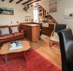A living room with a brown leather sofa a wooden coffee table a wooden kitchen with cabinets and a sink and chairs at Maple Tree Cottage in Longframlington