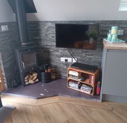 A corner with a wood burning stove firewood and tools a wall mounted TV and a small wooden shelf with books and DVDs at Silver Birch Lodge in Misterton