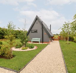 A gravel pathway leading to a small gray triangular house with a bench and surrounding trees and bushes at Silver Birch Lodge in Misterton