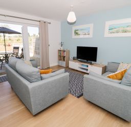 A living room with two gray sofas a TV on a stand a small bookcase and sliding doors leading to a patio with a table and chairs at 14 Parc Delfryn in Brynteg near Benllech