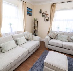 A living room with two sofas, a footstool, wall shelves with plants, and windows with curtains at Neland View in Windermere