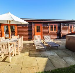 A patio area with wooden dining table and chairs under umbrella two wooden loungers and a hot tub outside a log cabin at Elm Lodge in Greetham Rutland