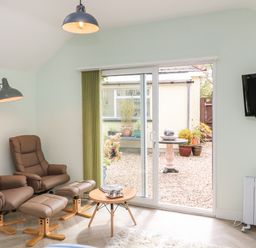 A small sitting area with two brown armchairs and matching footstools a small table a floor lamp a wall-mounted TV and sliding glass doors leading to a garden at Byghan Thornton in Illogan