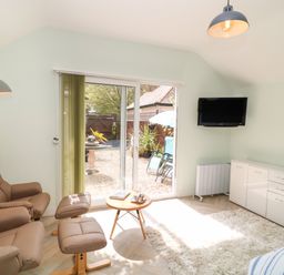 A room with beige armchairs a small round table a white cabinet a wall-mounted TV and glass sliding doors leading outside at Byghan Thornton in Illogan