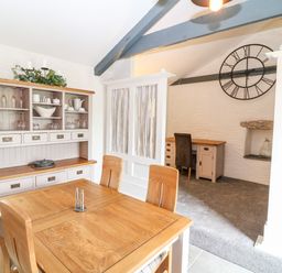 A dining area with a wooden table and chairs and a cabinet with glassware next to a carpeted study space with a desk chair and large wall clock at Polgear View Polgear Nr Redruth