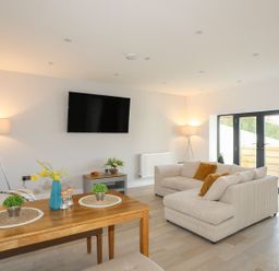 A living room with a beige sofa and yellow pillows next to glass doors and a wooden dining table with plants at Bryn Min Manton Bach in Caernarfon