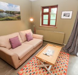 A living room with a beige sofa with purple cushions a wooden coffee table a patterned rug a floor lamp two framed pictures on the wall and windows at Bwthyn Canol in Haverfordwest