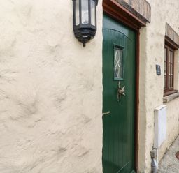 A close-up of a green door with a decorative knocker set in a cream textured wall with a window and outdoor light at Bwthyn Canol in Haverfordwest
