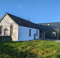 A white stone building with three arched windows and a purple door attached to a modern dark gray building at Ysgoldy Brynwyre in Llanrhystud