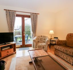 A living room with a TV on a wooden stand a patterned armchair a brown sofa a glass coffee table and a lamp by the sofa at Friars Fold in Newby Bridge