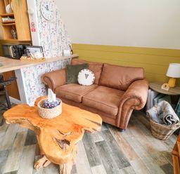 A living area with a brown sofa, a wooden coffee table, a small side table with a lamp, a basket with blankets, and a kitchen counter with chairs at Buttercup Lodge in Misterton
