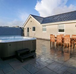 A patio with a hot tub and wooden dining table with chairs at Yr Hen Feudy in Moelfre