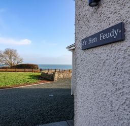 View of a stone house exterior wall with a sign Yr Hen Feudy and a fenced grassy yard with a tree and ocean in background at Yr Hen Feudy in Moelfre