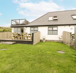 An outdoor patio with wooden table and benches adjacent to a house with fenced grass yard at Bwthyn Awel in Church Bay near Cemaes Bay