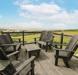 A wooden deck with five wooden chairs and a small table overlooking a field at Bwthyn Awel in Church Bay near Cemaes Bay