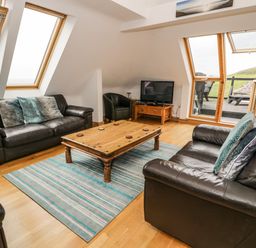 A living room with two black leather sofas a wooden coffee table a patterned blue rug a television on a wooden stand and large windows at Bwthyn Awel in Church Bay near Cemaes Bay