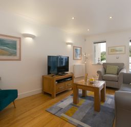 A living room with gray and green chairs a wooden table with drinks and snacks and a TV on a wooden stand at The Waves in St Ives
