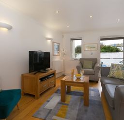 A living room with a television on a wooden stand a coffee table with snacks and drinks a grey armchair a grey sofa and a green chair at The Waves in St Ives