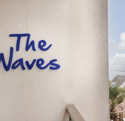 A white wall with the words The Waves in blue with a railing and a view of a building and the sea at The Waves in St Ives