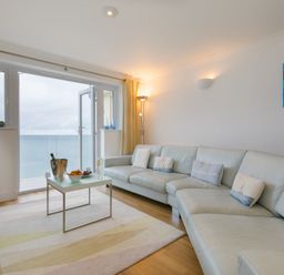 A living room with a large sofa coffee table and an open door leading to a balcony with a sea view at Turquarze in Carbis Bay