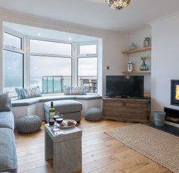 A living room with a grey sofa, bay window seat, TV on a wooden chest, fireplace, and wooden floor at Whitsunday House in Carbis Bay