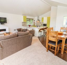 An open living space with brown sofas a wooden dining table with chairs a kitchen island with bar stools and a wall mounted television at Oak Lodge in Glasshouse